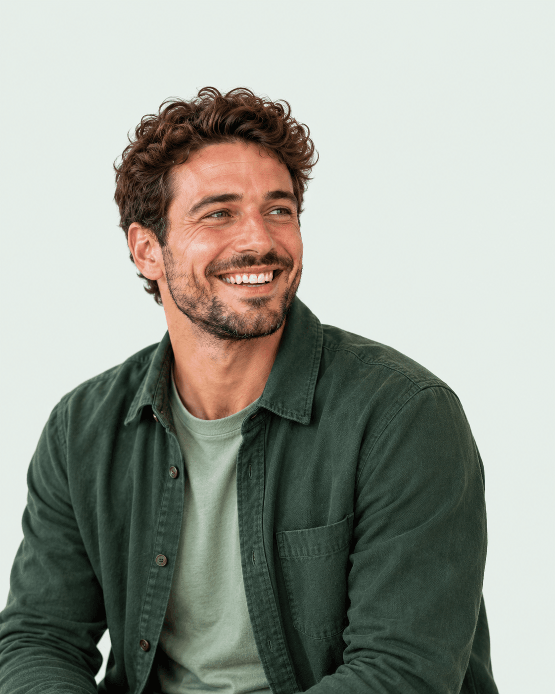 A smiling man with curly hair in a dark green overshirt, looking to one side, photographed on a plain pale-green background.